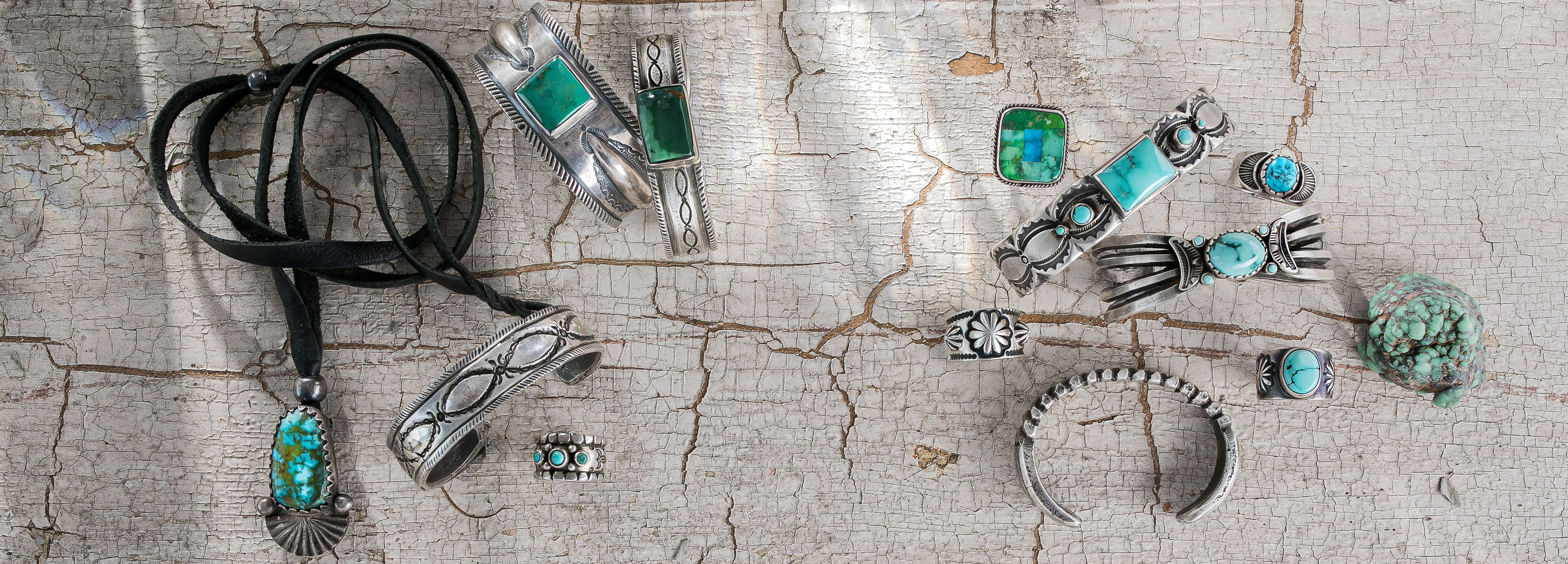 Various pieces of silver and turquoise jewelry in necklaces, bands, and rings sit on a cracked white surface.